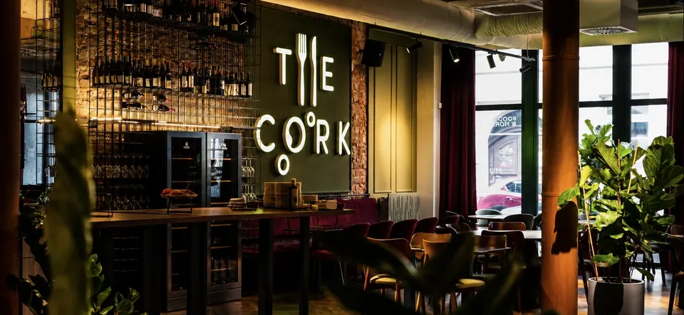 Sala The Cork przy Rzeźniczej: podświetlony napis THE CORK na ciemnozielonej ścianie, regał z winami, wysoki stół i okna na ulicę